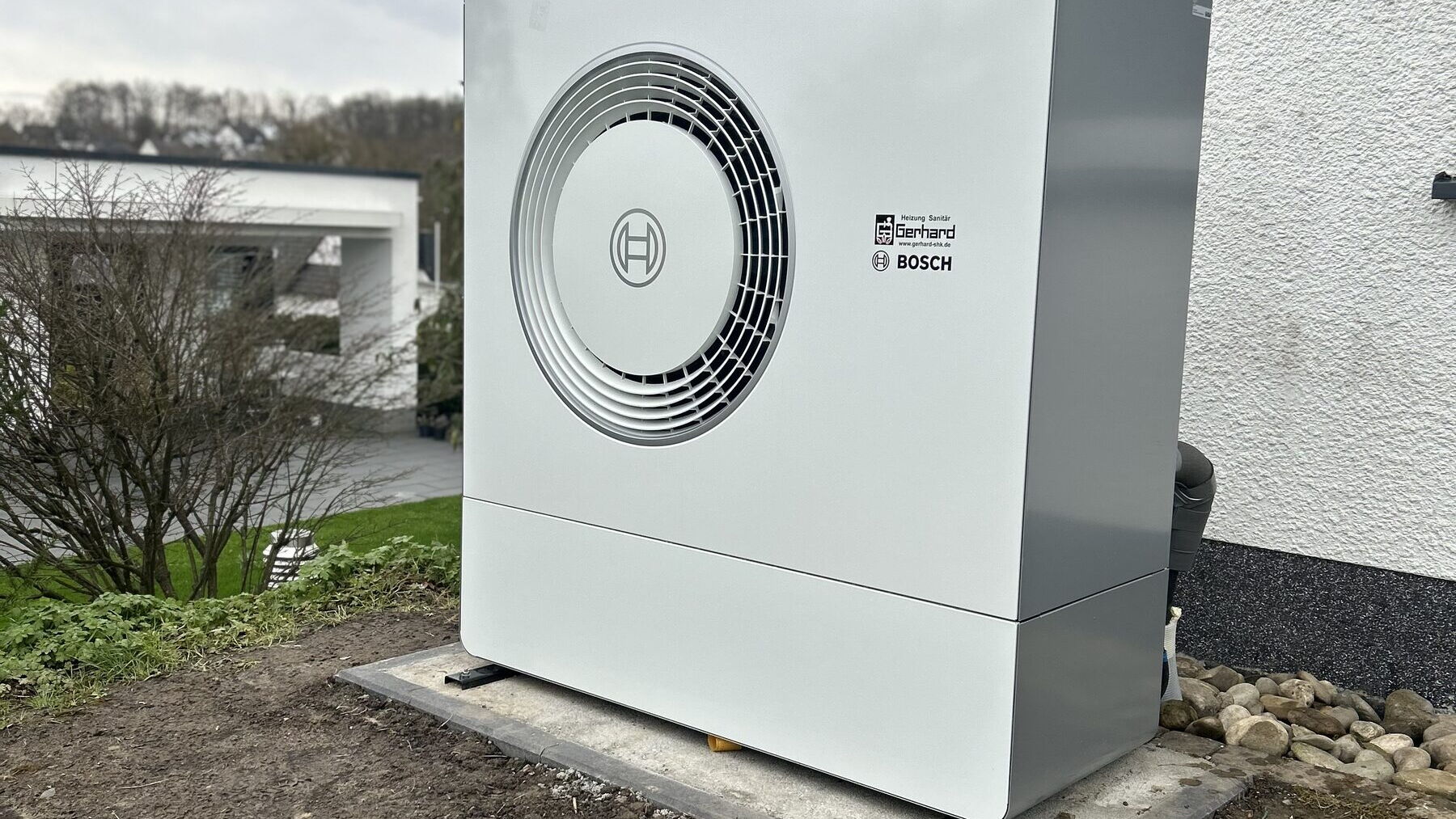 Weiße Bosch-Wärmepumpe auf Betonfundament neben einem verputzten Einfamilienhaus